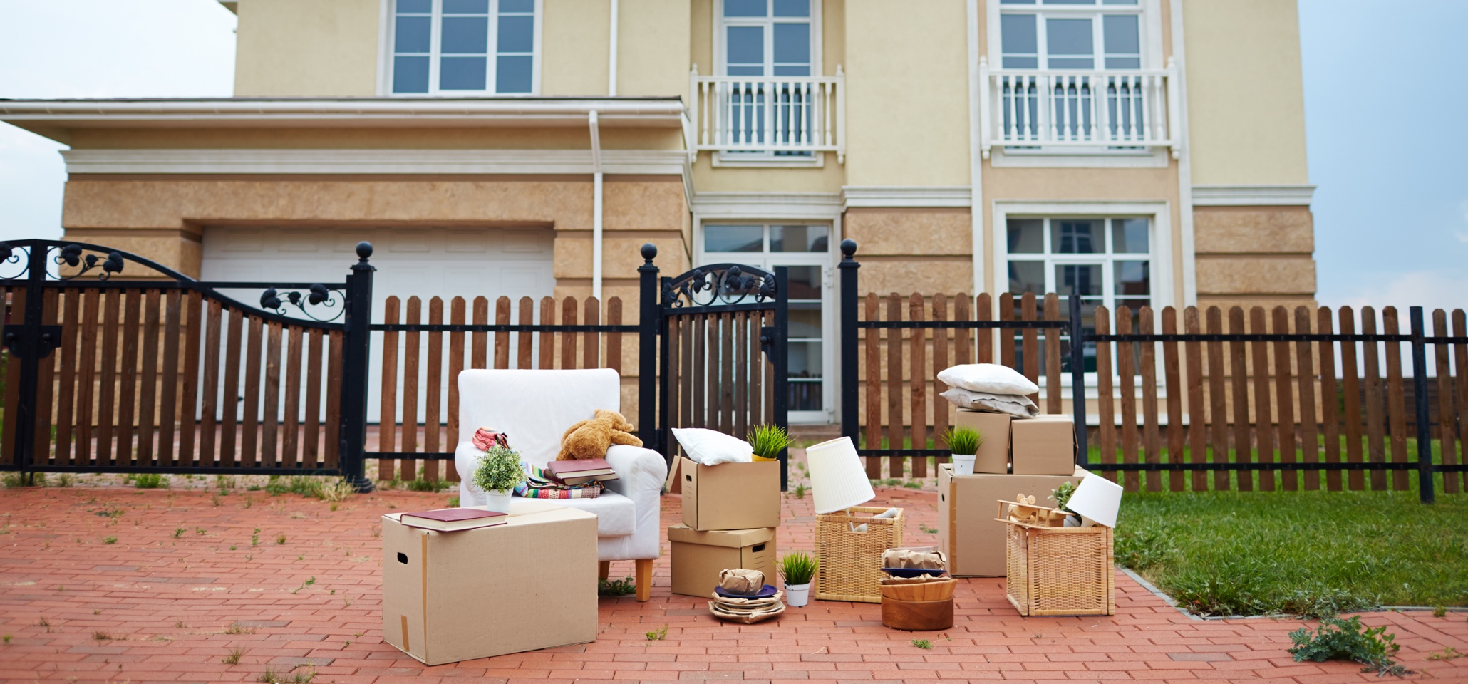 boxes and furniture on a red brick driveway outside a large house with a low fence